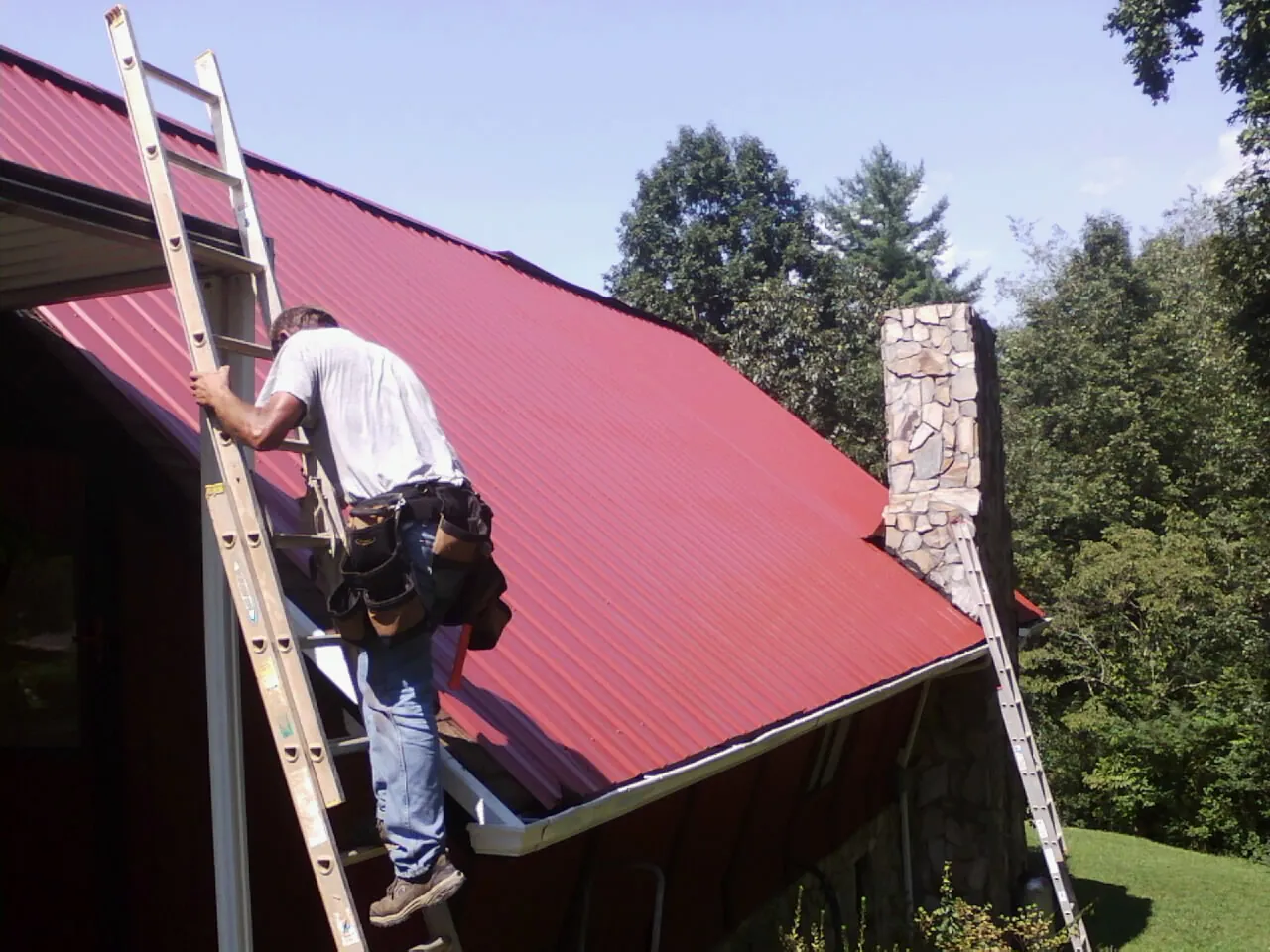 Roofer on a ladder beside a red metal roof and stone chimney — Soffit & Fascia Repair by Roofing Brothers in Dodge